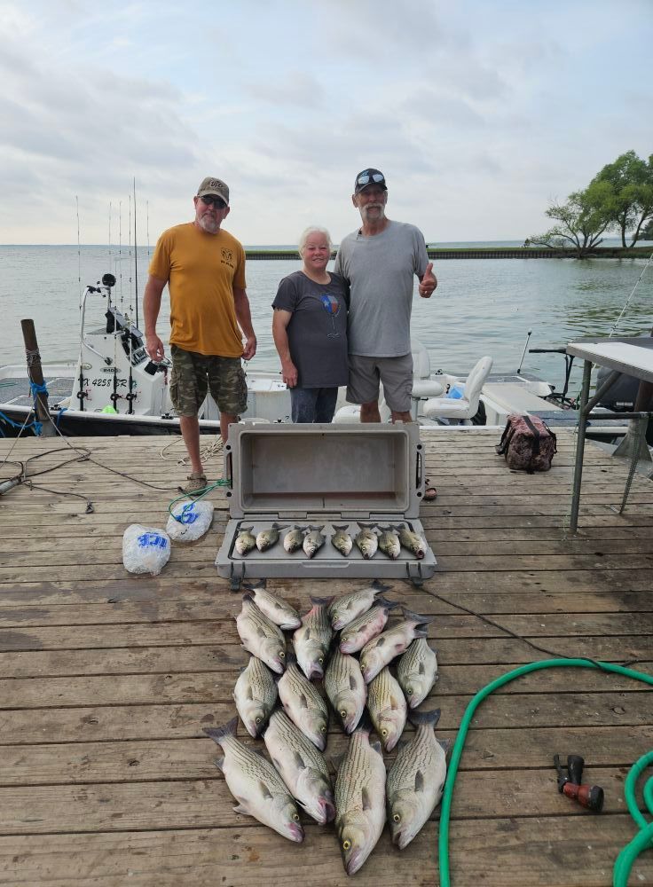 Three people stand on a wooden dock next to a boat, a large cooler, and a large catch of fish on the planks.