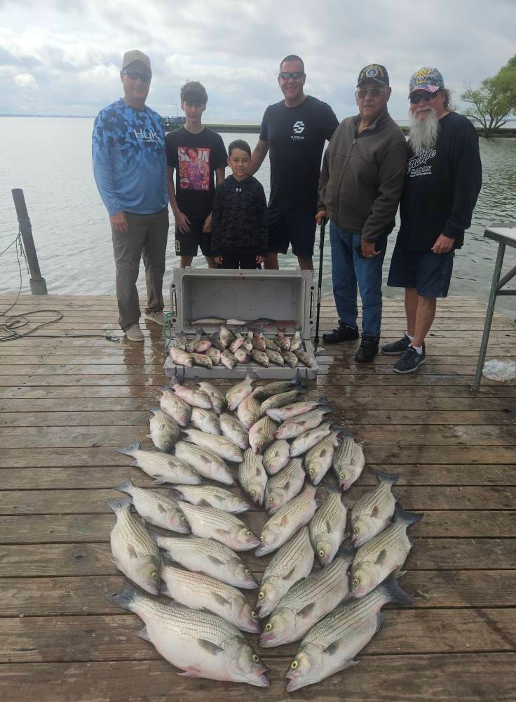 Six people pose on a wooden dock with a large catch of fish spread out on the planks and inside a cooler.