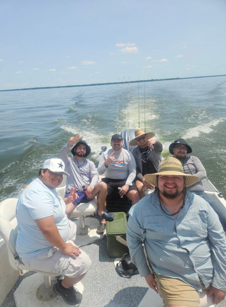 Five people riding in a boat on the water on a sunny day, with a fishing rod in the background and boat wake behind them.