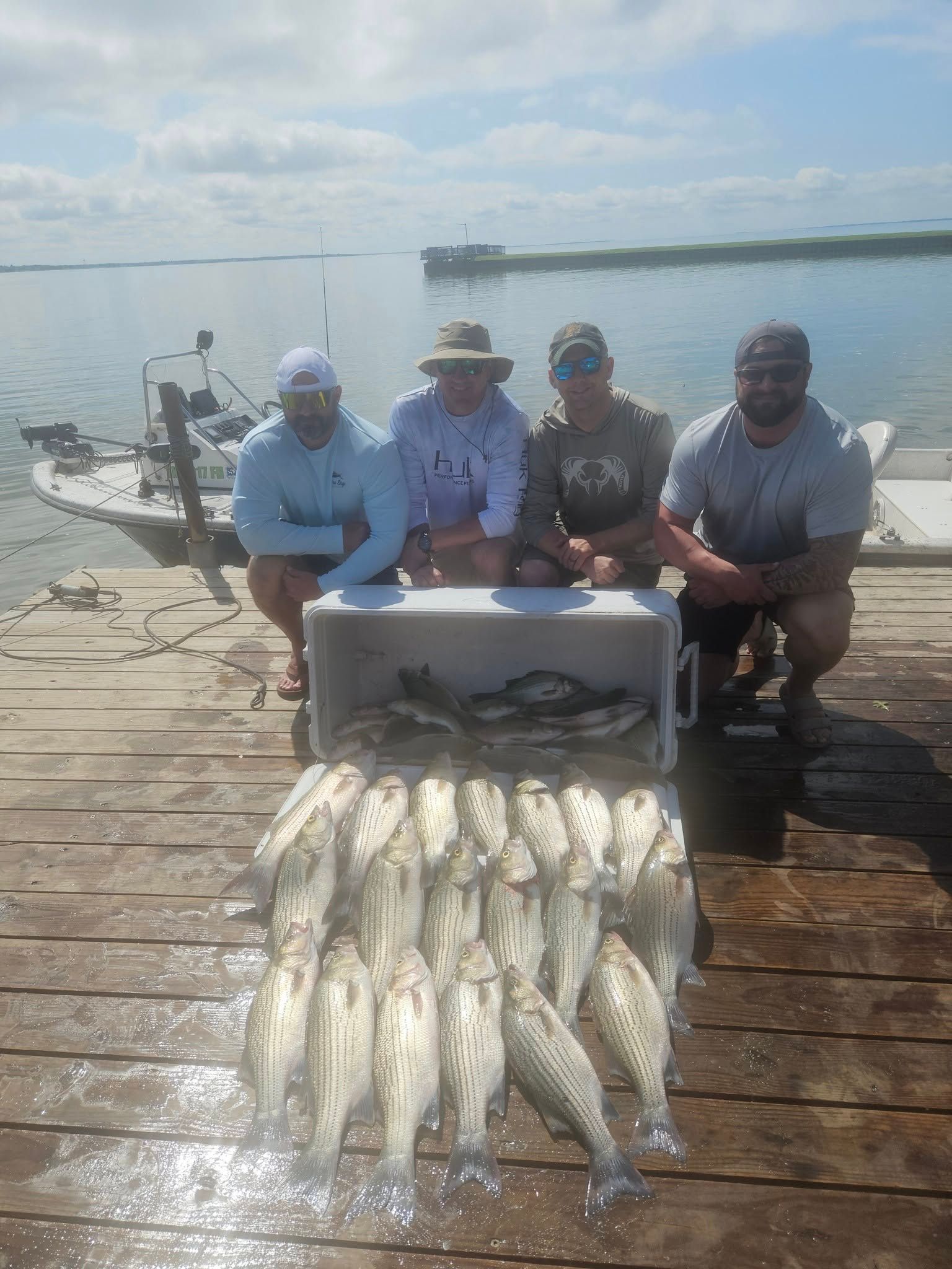 Four people pose on a wooden dock with a cooler full of fish and a boat in the background on a sunny day.