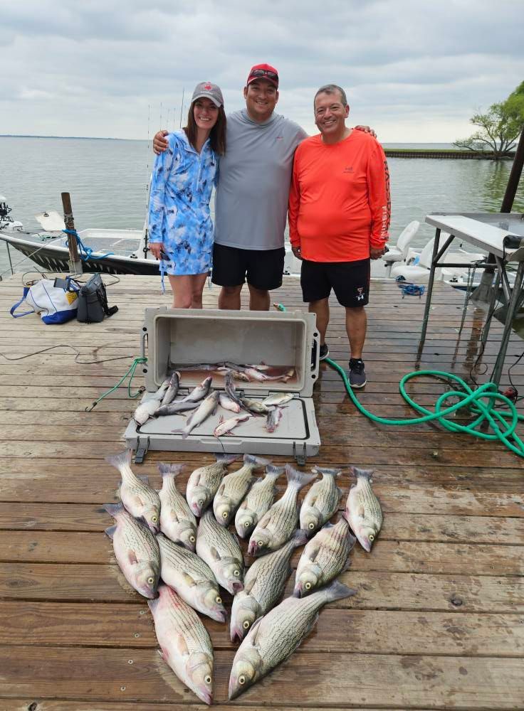 Three people stand on a wooden dock with a large cooler of fish and a display of fresh catch arranged on the deck.