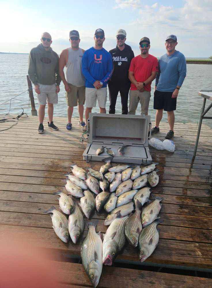 A group of six men stand on a wooden dock behind a large cooler filled with their catch of striped bass.