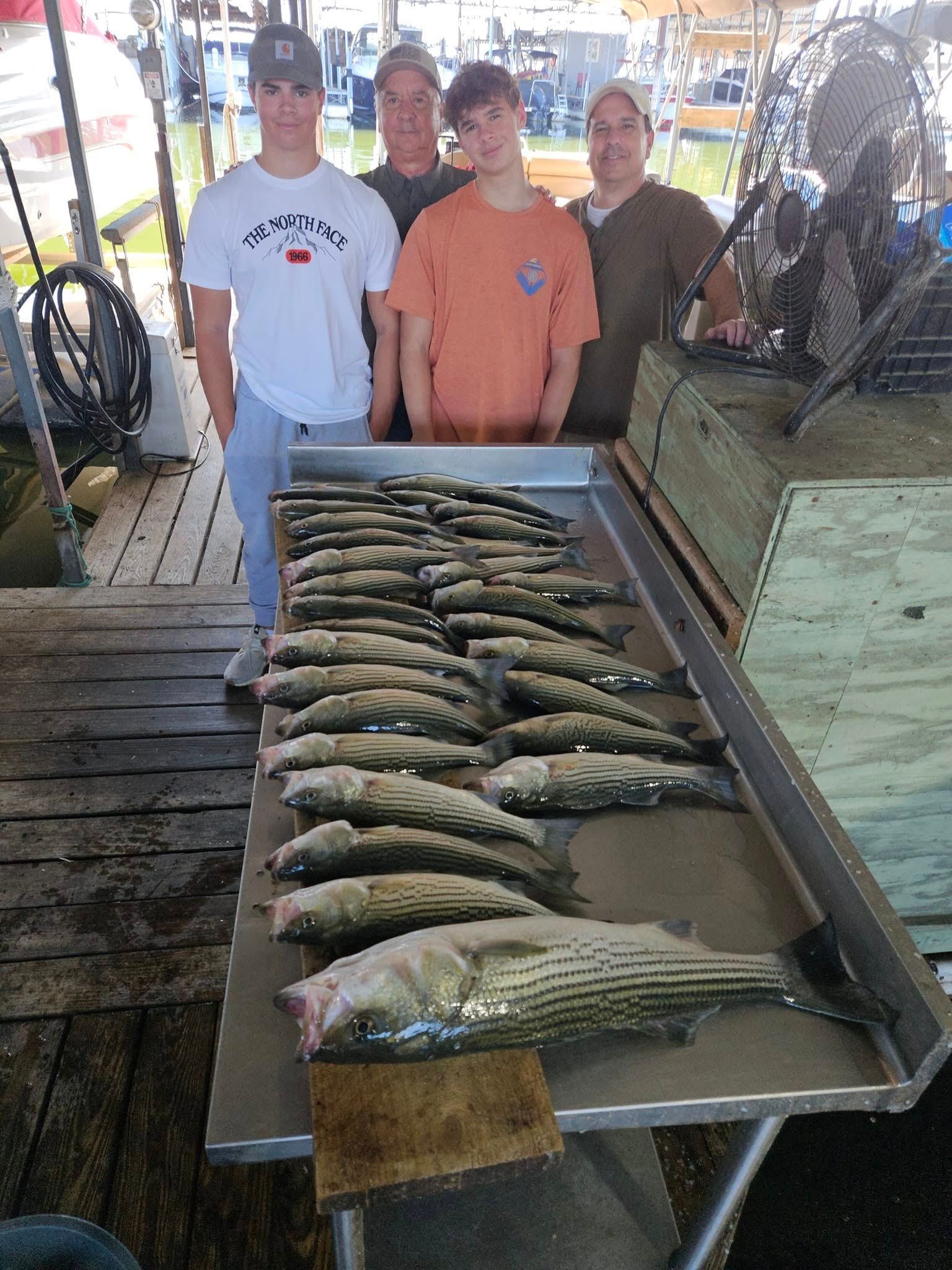 Four people stand behind a table covered with a large catch of striped fish at a dock.