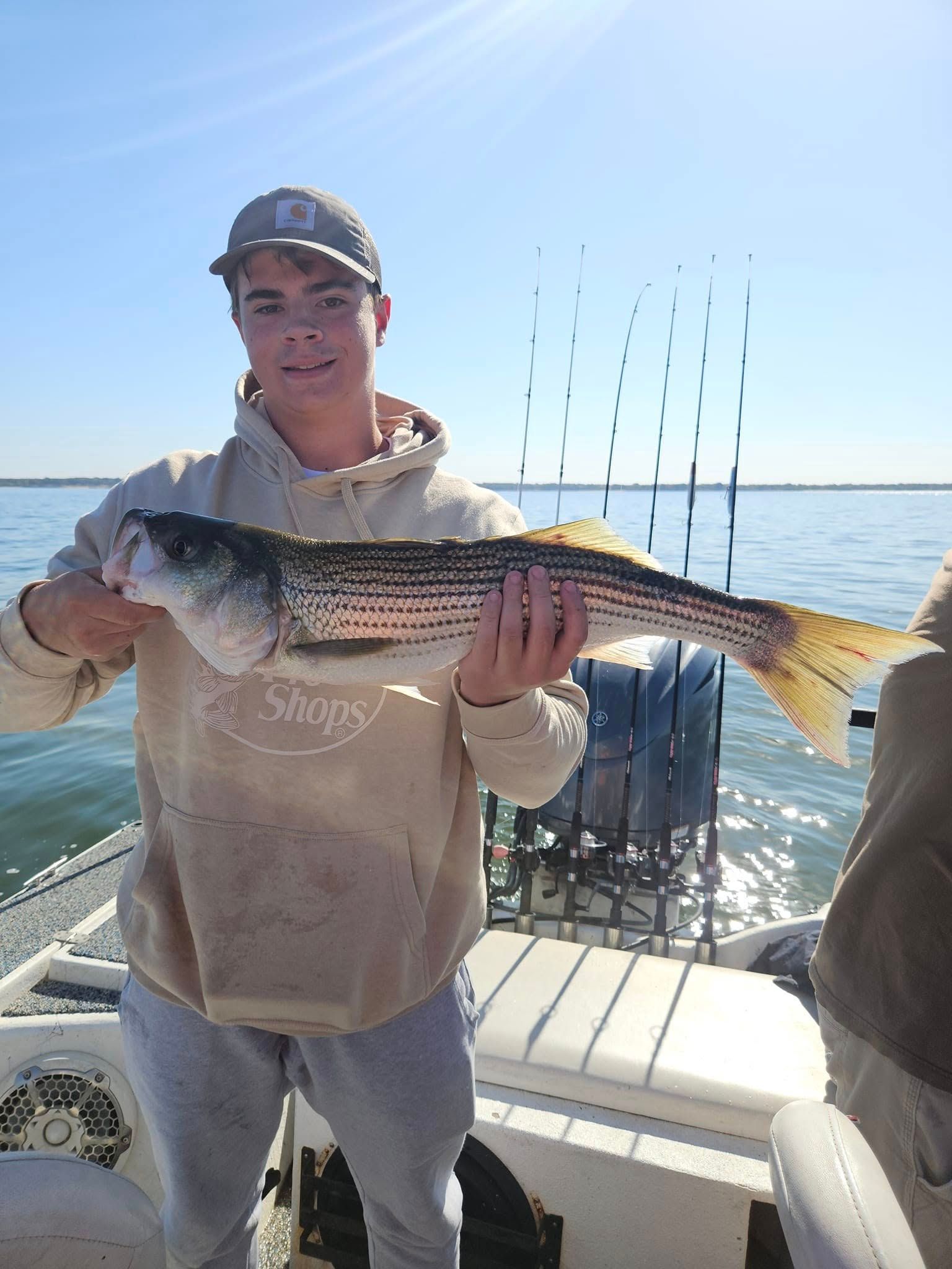A person in a tan hoodie holding a striped bass on a boat, with fishing rods in the background under a clear sky.