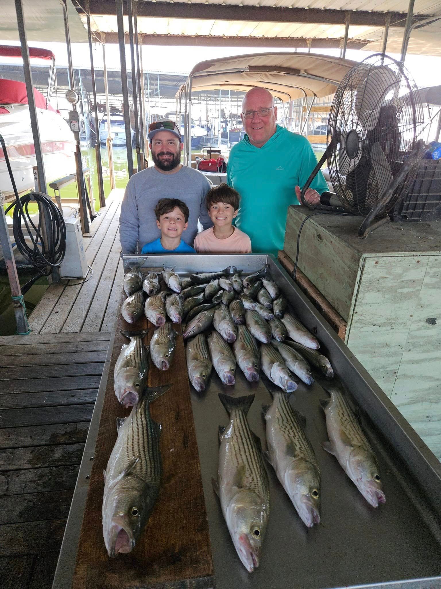Two adults and two children stand behind a stainless steel table filled with a large catch of striped fish at a dock.
