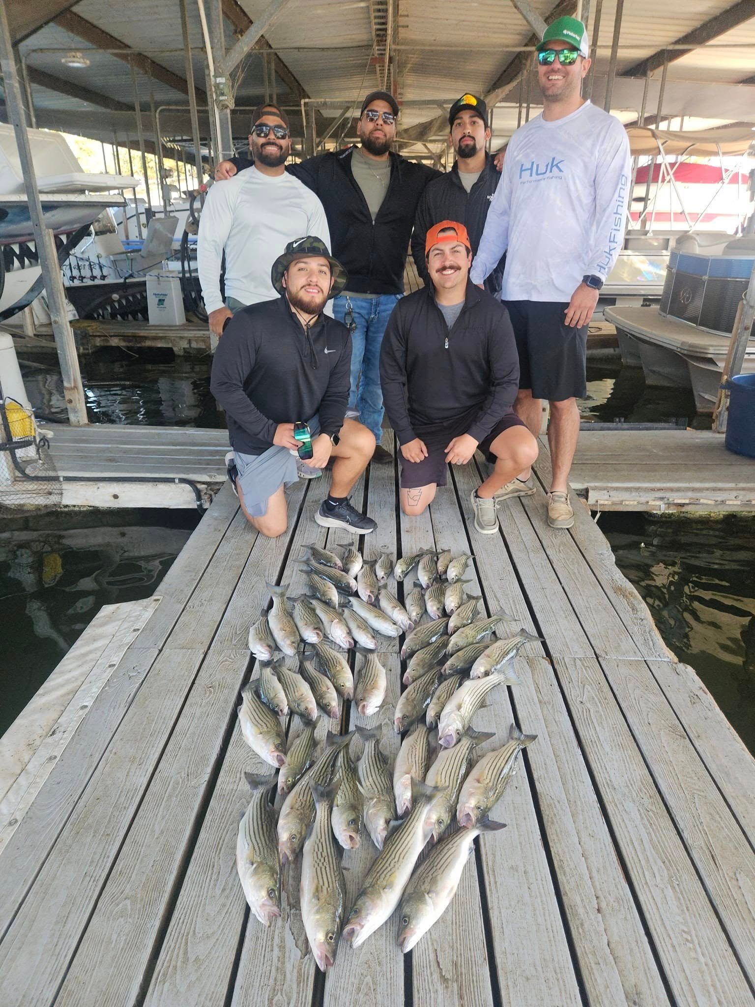 A group of seven people posing on a wooden dock with a large catch of fish spread out on the ground in front of them.