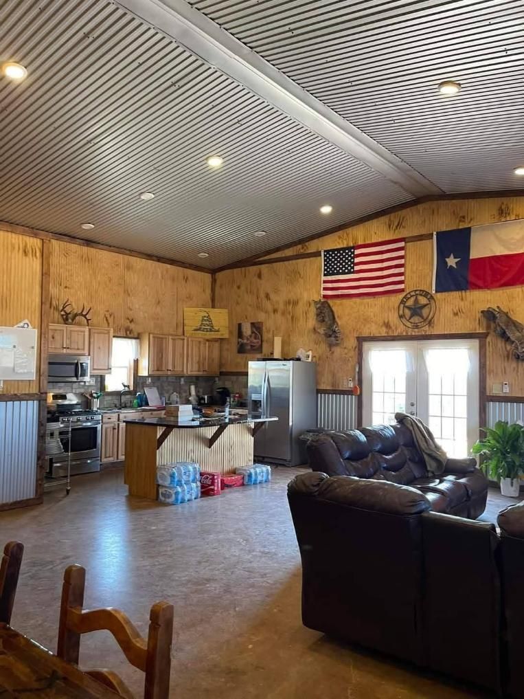 A spacious, rustic living area with plywood walls, a metal ceiling, a kitchen, and a dark leather sofa.