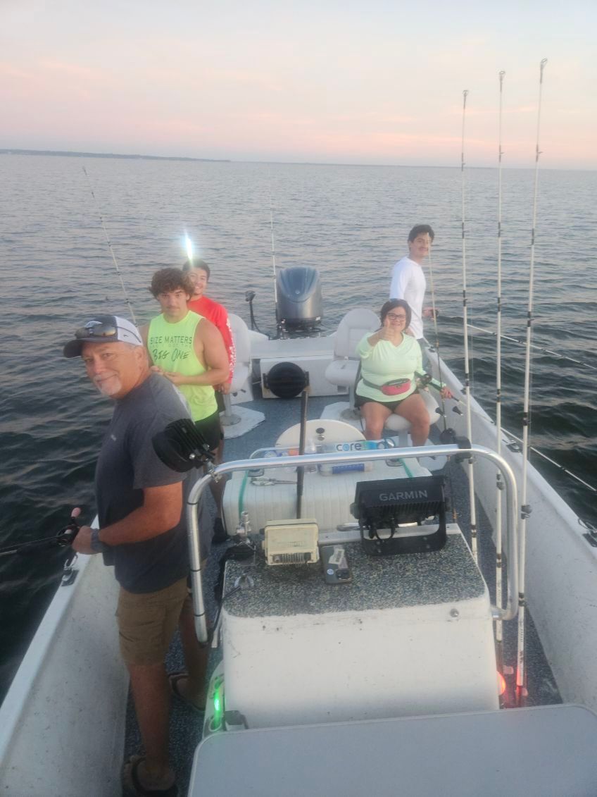 Four people on a fishing boat on the water at sunset, with fishing rods in holders and a console in the foreground.