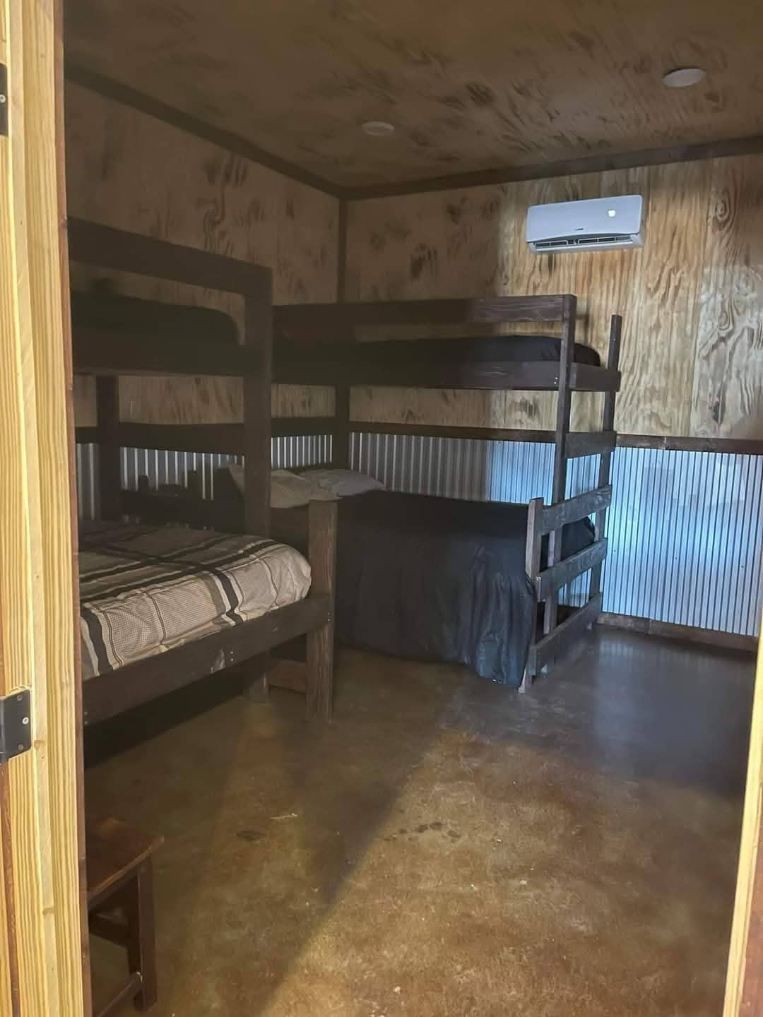 A small room with plywood walls, a concrete floor, two bunk beds, and a wall-mounted air conditioner.