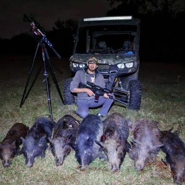 A person sits in front of a utility vehicle in a field at night, holding a rifle behind seven wild hogs on the grass.