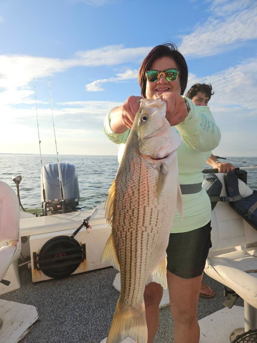 A person holding a large striped bass on a boat, with the ocean in the background.