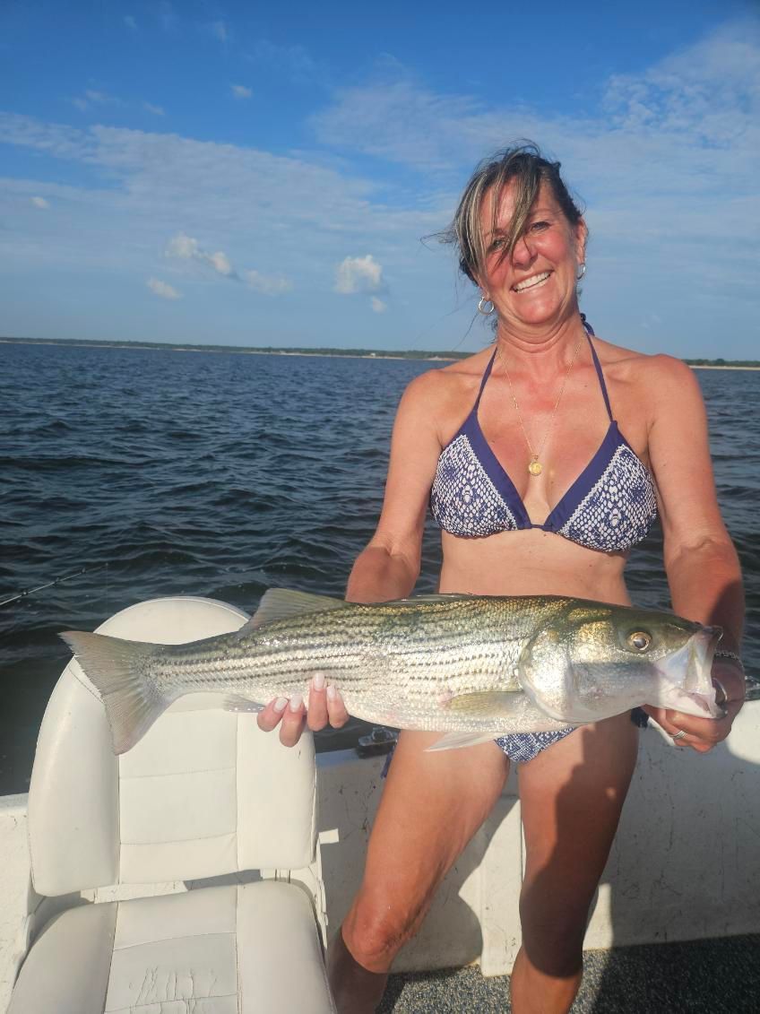 A person in a bikini smiles while holding a large, striped fish on a boat on the water under a blue sky.