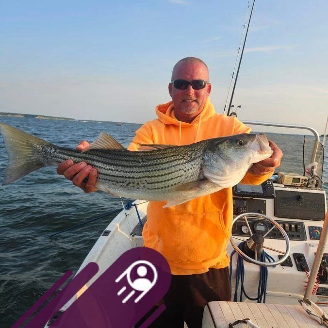 A person wearing a bright orange hoodie and sunglasses holds a large striped bass while standing on a boat.