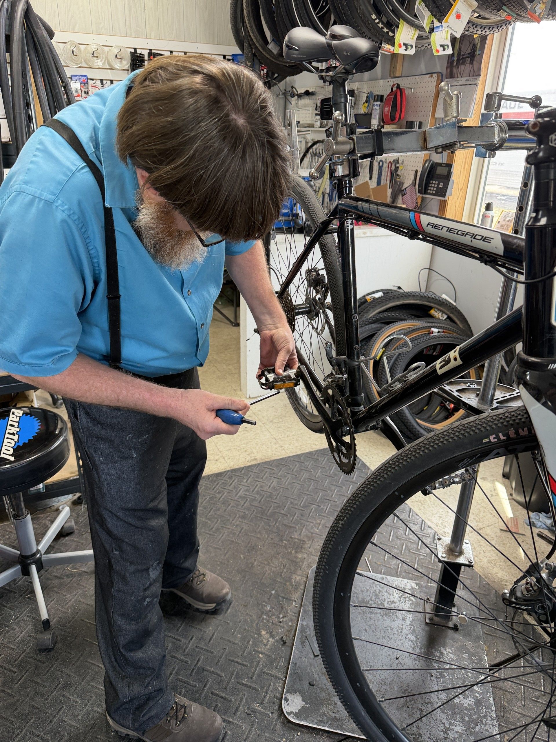 A person in a blue shirt and suspenders uses a screwdriver to work on a bicycle mounted on a stand in a workshop.