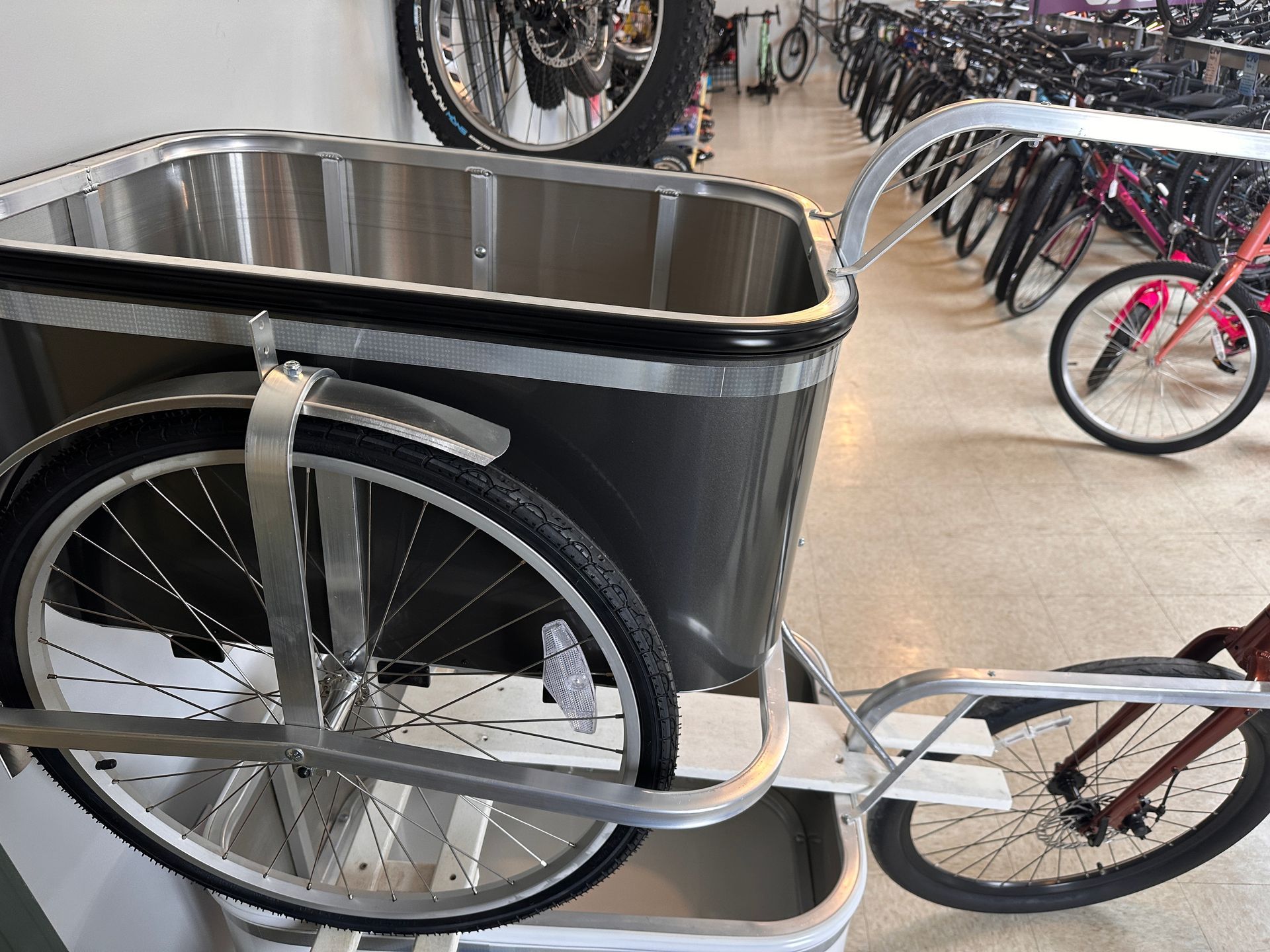 A cargo bike with a silver box-like container, parked inside a bike shop. Black tires and a gray frame are visible.