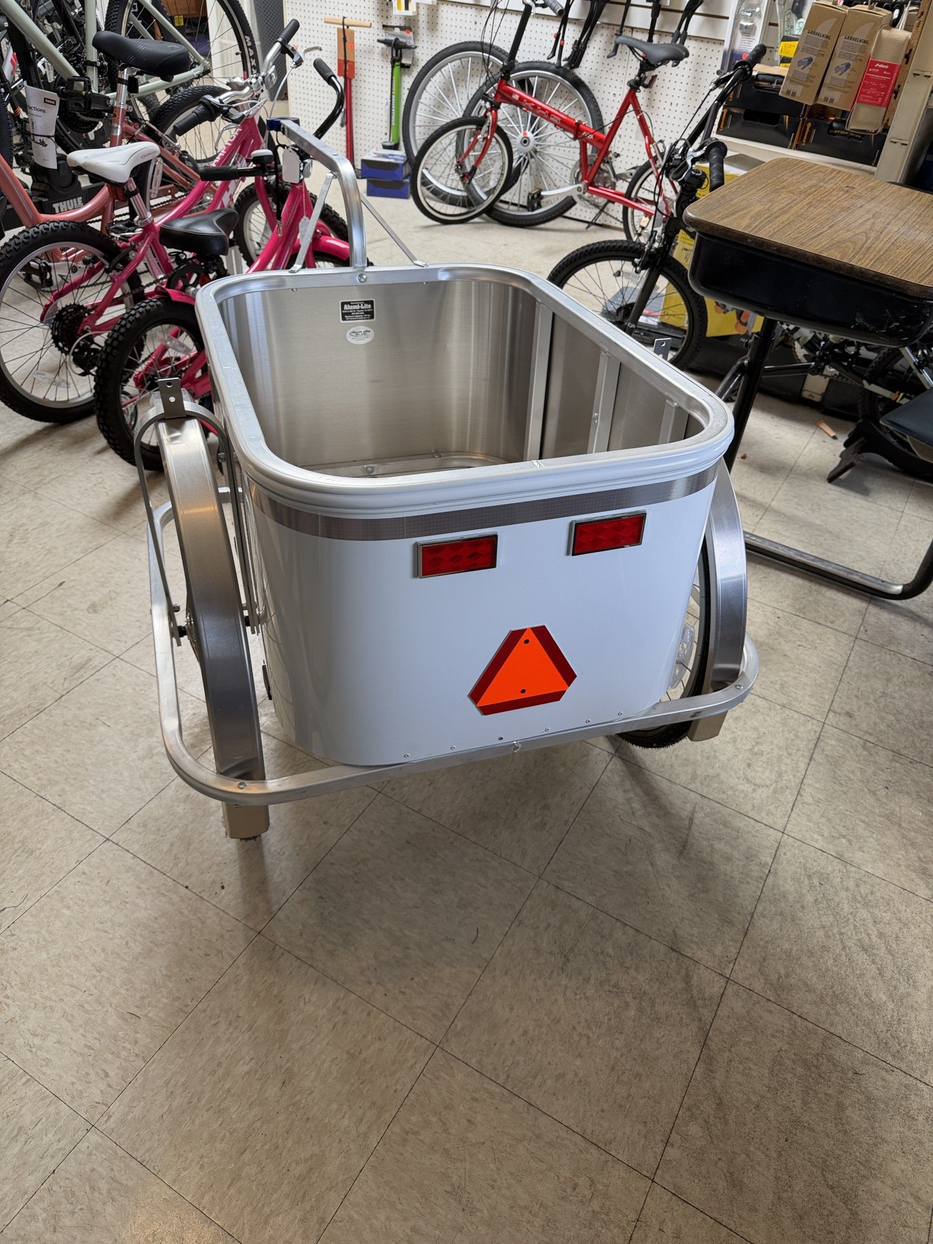 A silver bicycle cargo trailer with a deep, rectangular bin and red reflective safety triangles on the rear.