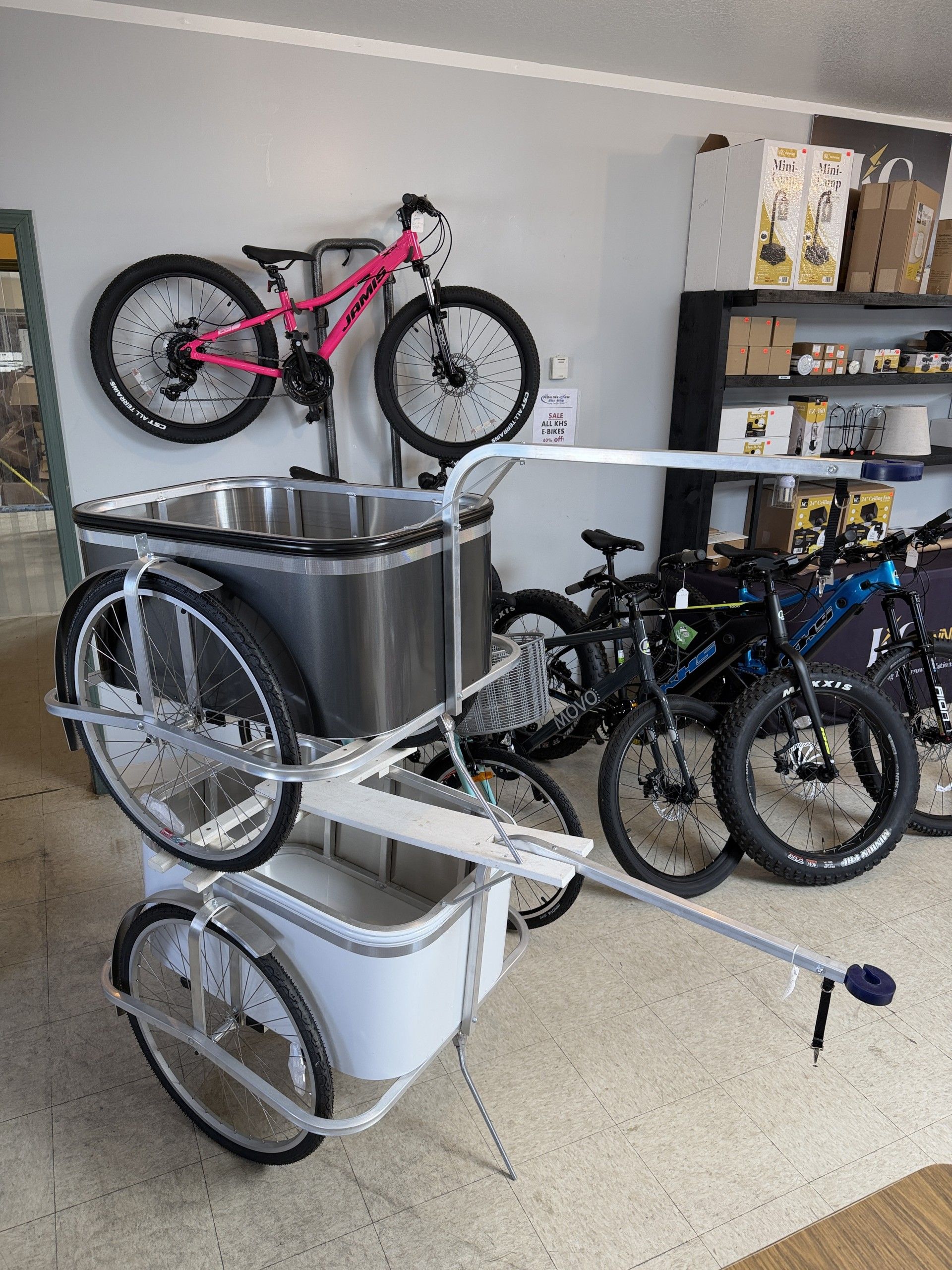 A dual-level bicycle trailer with silver and white bins, displayed in a shop with bicycles in the background.