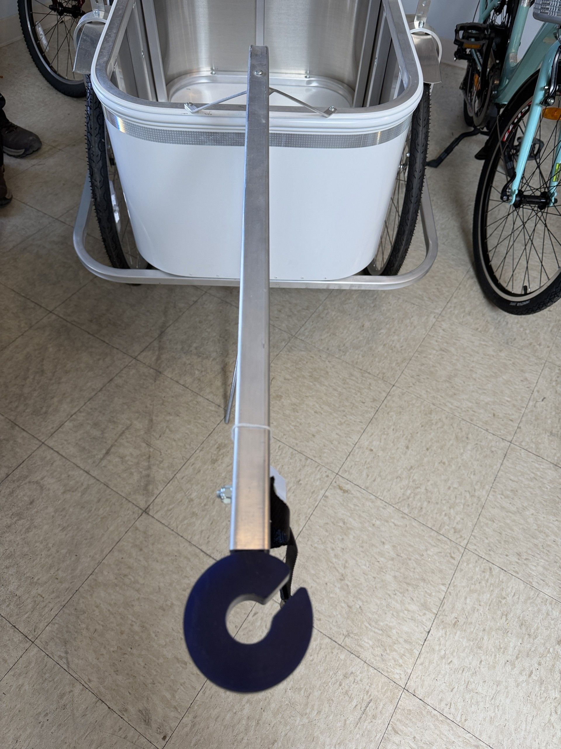 A white bicycle cargo trailer with a long silver towing arm extending toward a circular black hitch attachment.