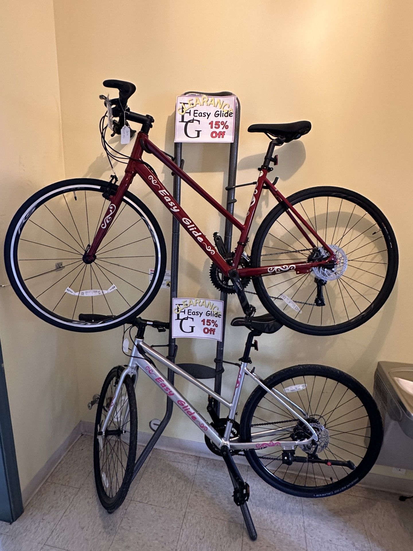 Two bicycles, a red one on top and a silver one below, are mounted on a metal rack against a tan wall. Both have price tags.