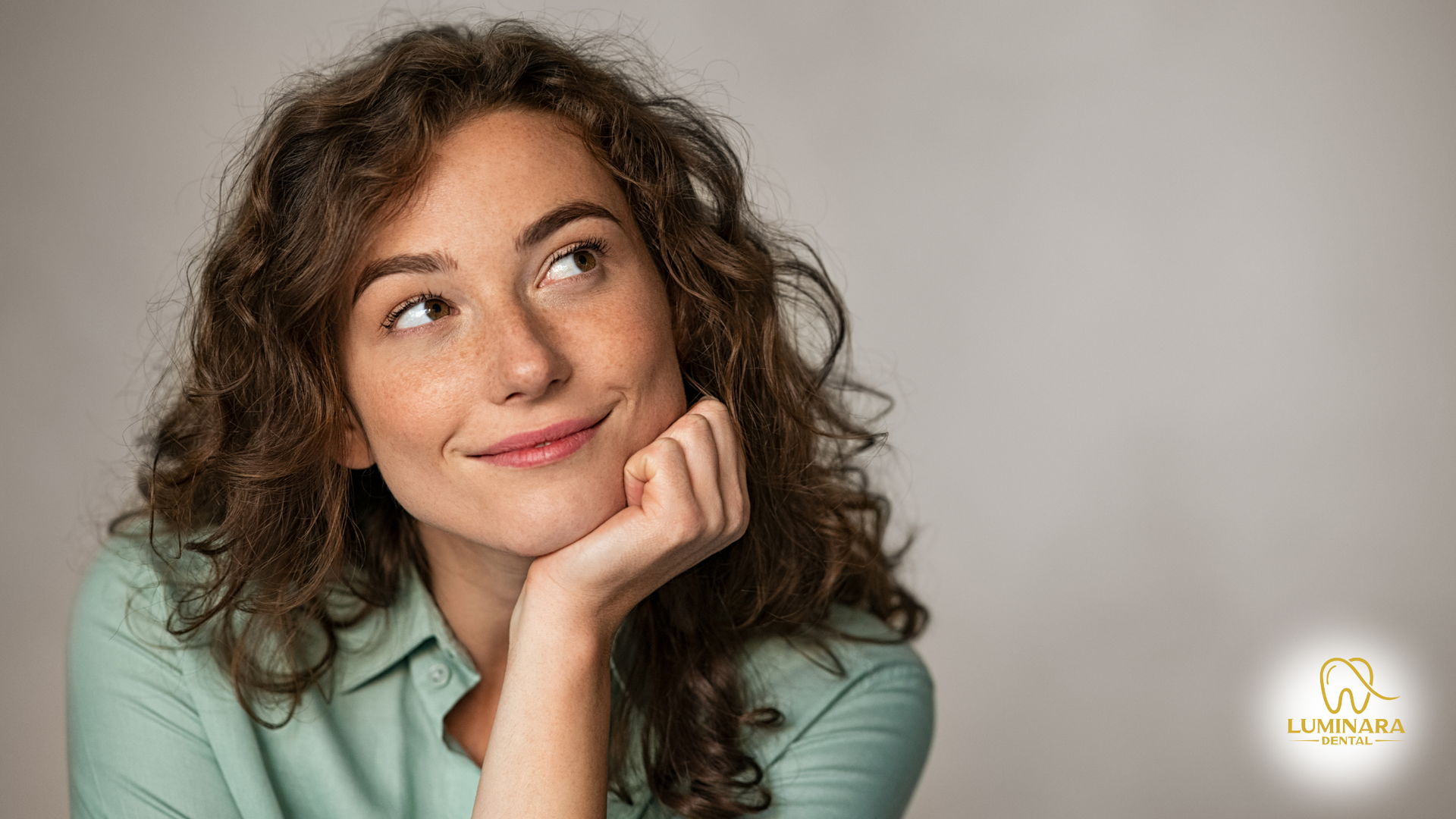 Woman with curly brown hair, smiling, looking upwards, resting chin on hand, against a light gray background.