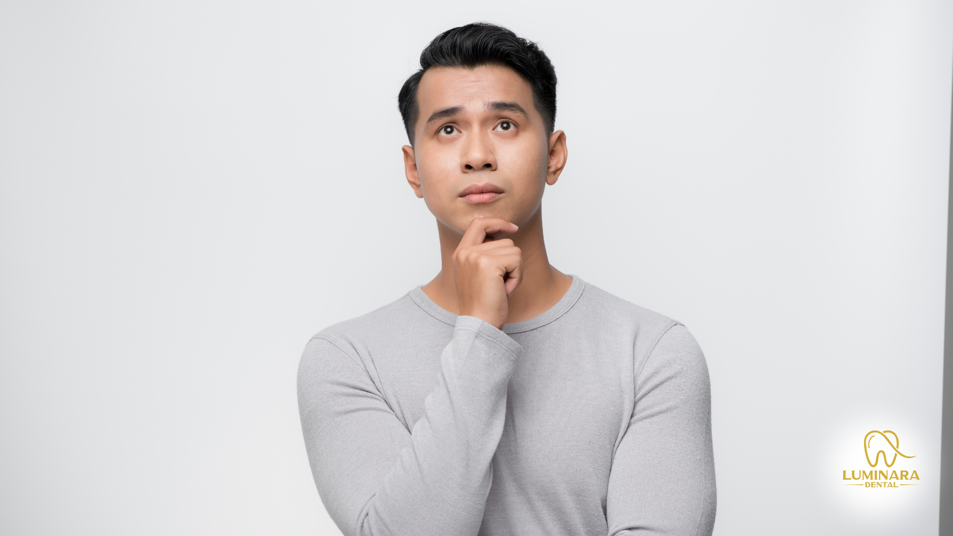 A man in a grey sweater is thinking with his hand on his chin.