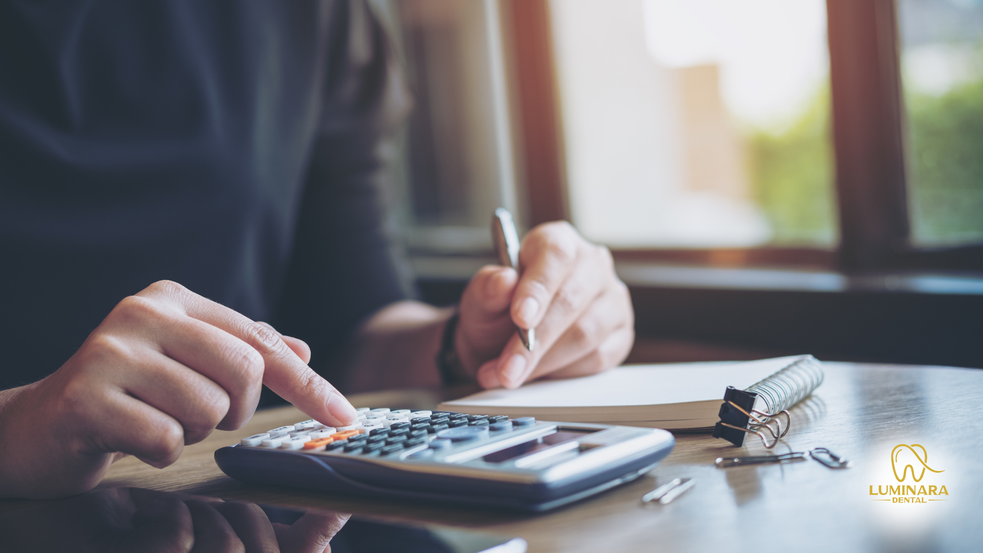 Person using a calculator with a pen and notepad nearby. Bright sunlight streams in from a window.