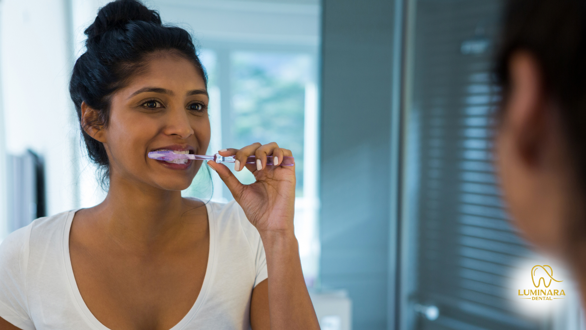 Woman brushing her teeth in front of a mirror, smiling. She's wearing a white shirt; bathroom setting.