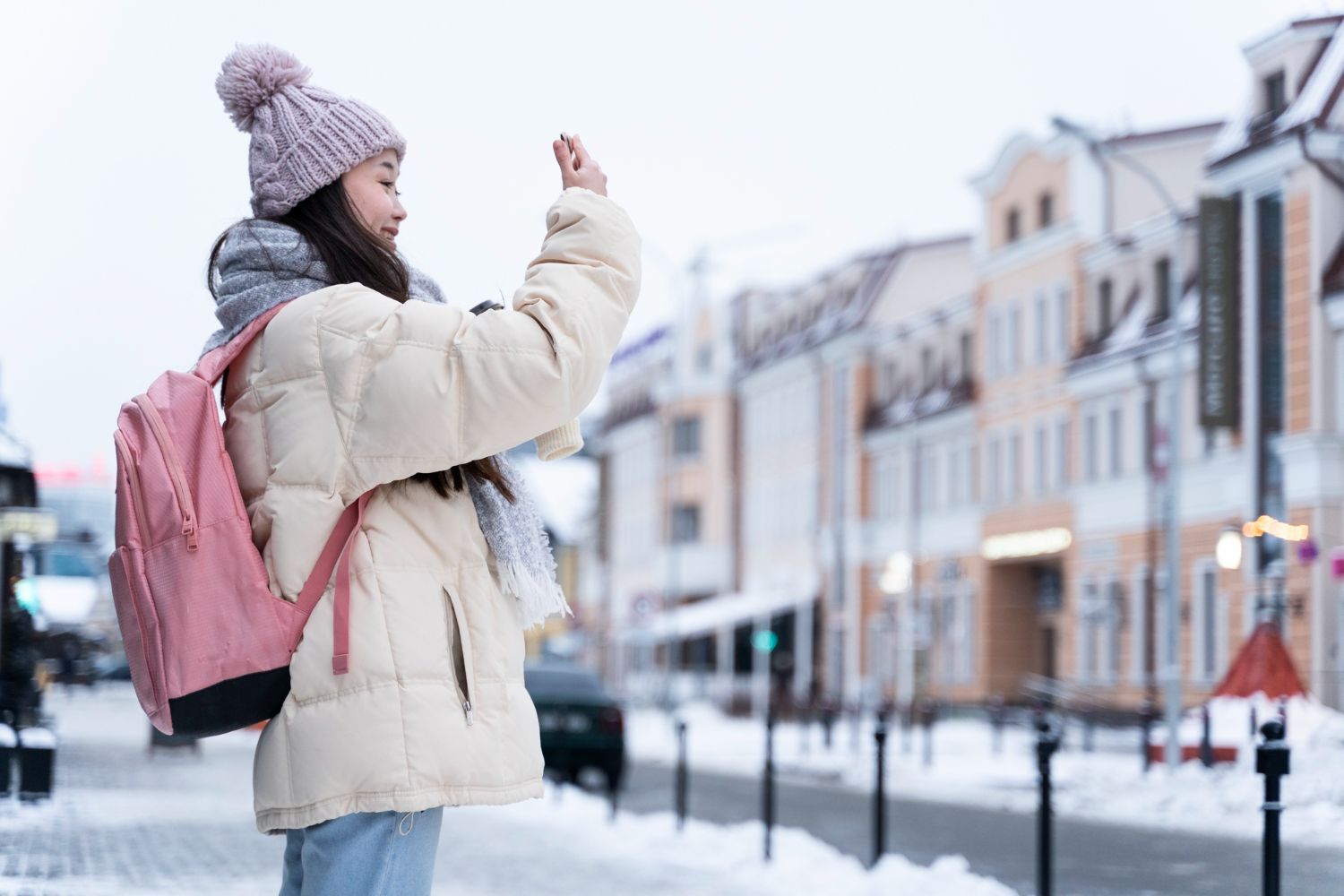 A young girl with a pink backpack is taking a picture of a snowy city street.