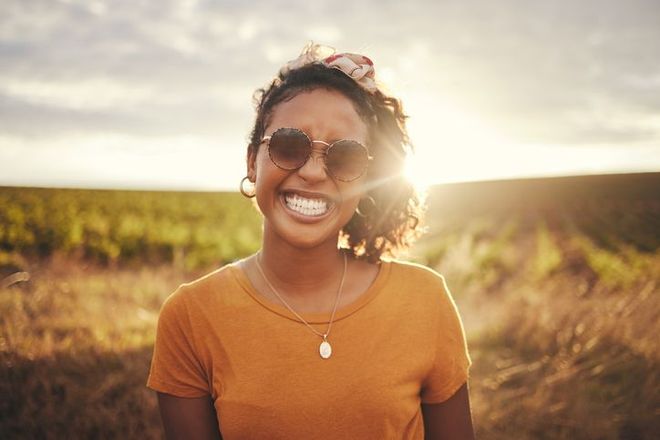 A woman wearing sunglasses is smiling in a field at sunset.