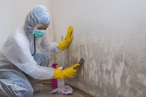 A woman in a protective suit is cleaning a wall with a spatula.