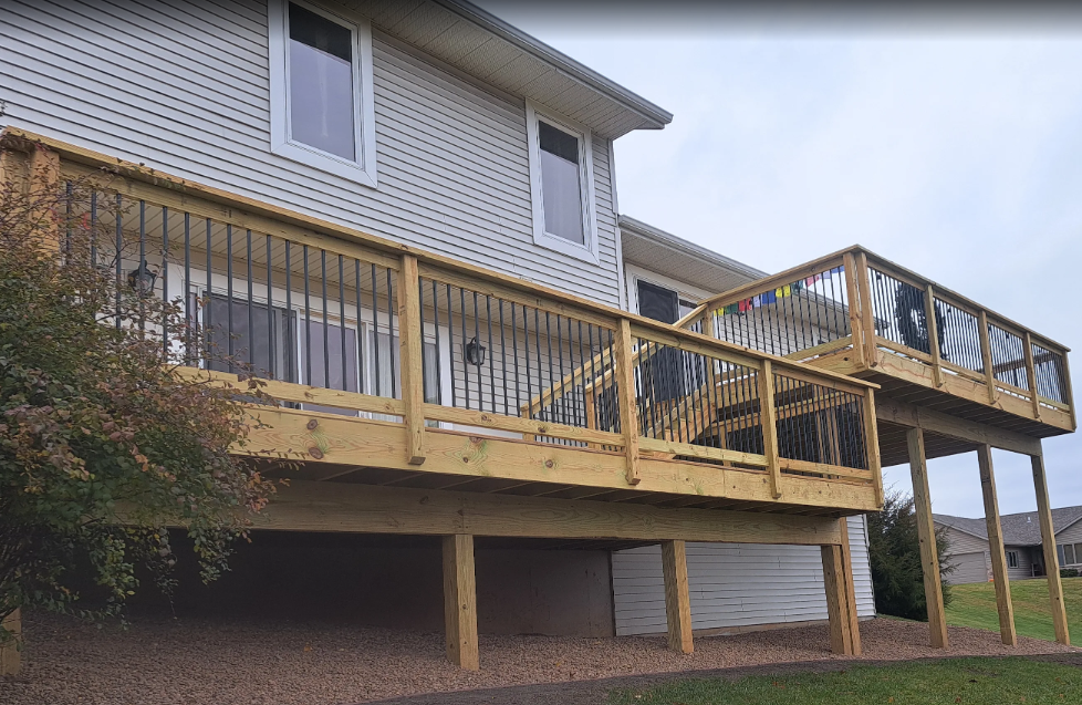 Two-story house with wooden decks. The lower deck has supporting posts. The railing is black and the deck is wooden.