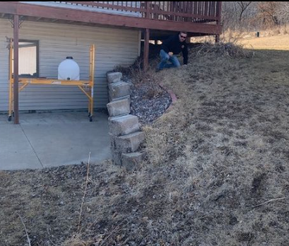 Two people near a house working on a slope. Brown grass, retaining wall, scaffolding, and a deck are visible.