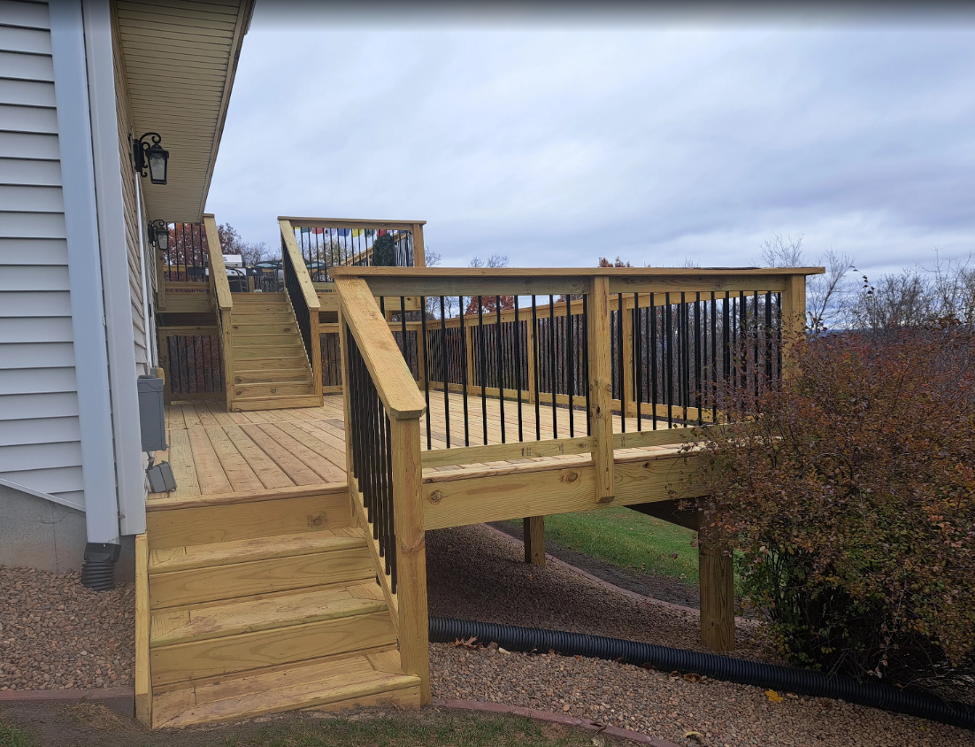 Wooden deck with stairs attached to a house with black railing. Overcast sky.