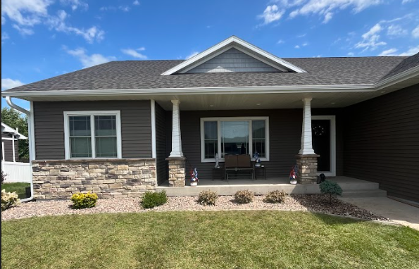 A house with brown siding, white columns, and a front porch under a blue sky. Landscaping in front with flowers and a stone facade.