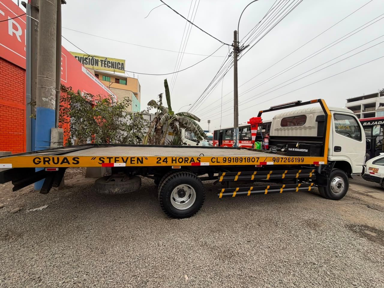 Una grúa transporta un camión blanco en una calle junto a un edificio y postes de servicios públicos.