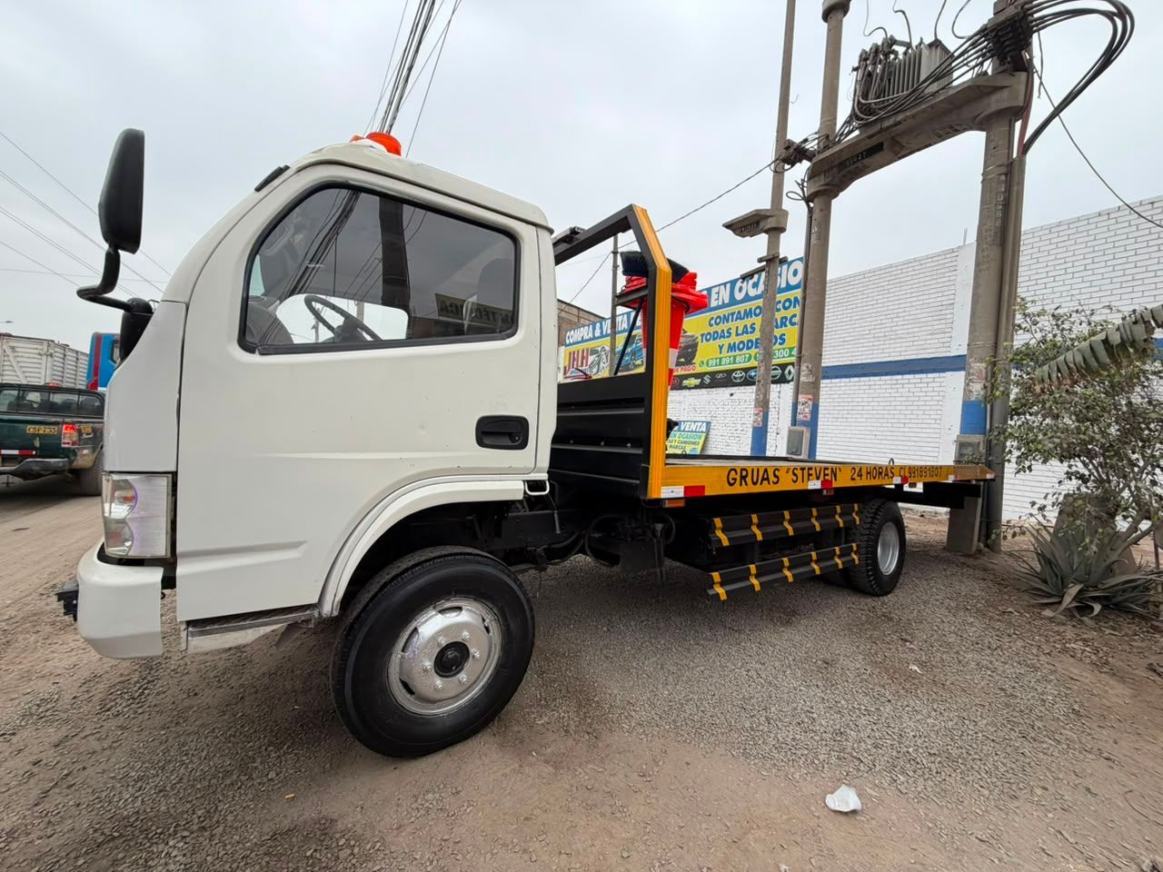 Camión blanco de plataforma plana estacionado en un terreno de tierra, vista lateral con postes de servicios públicos y edificios al fondo.