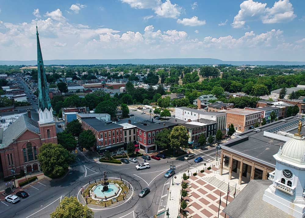 Aerial view of a town square with a church steeple, roundabout, and buildings under a blue sky.