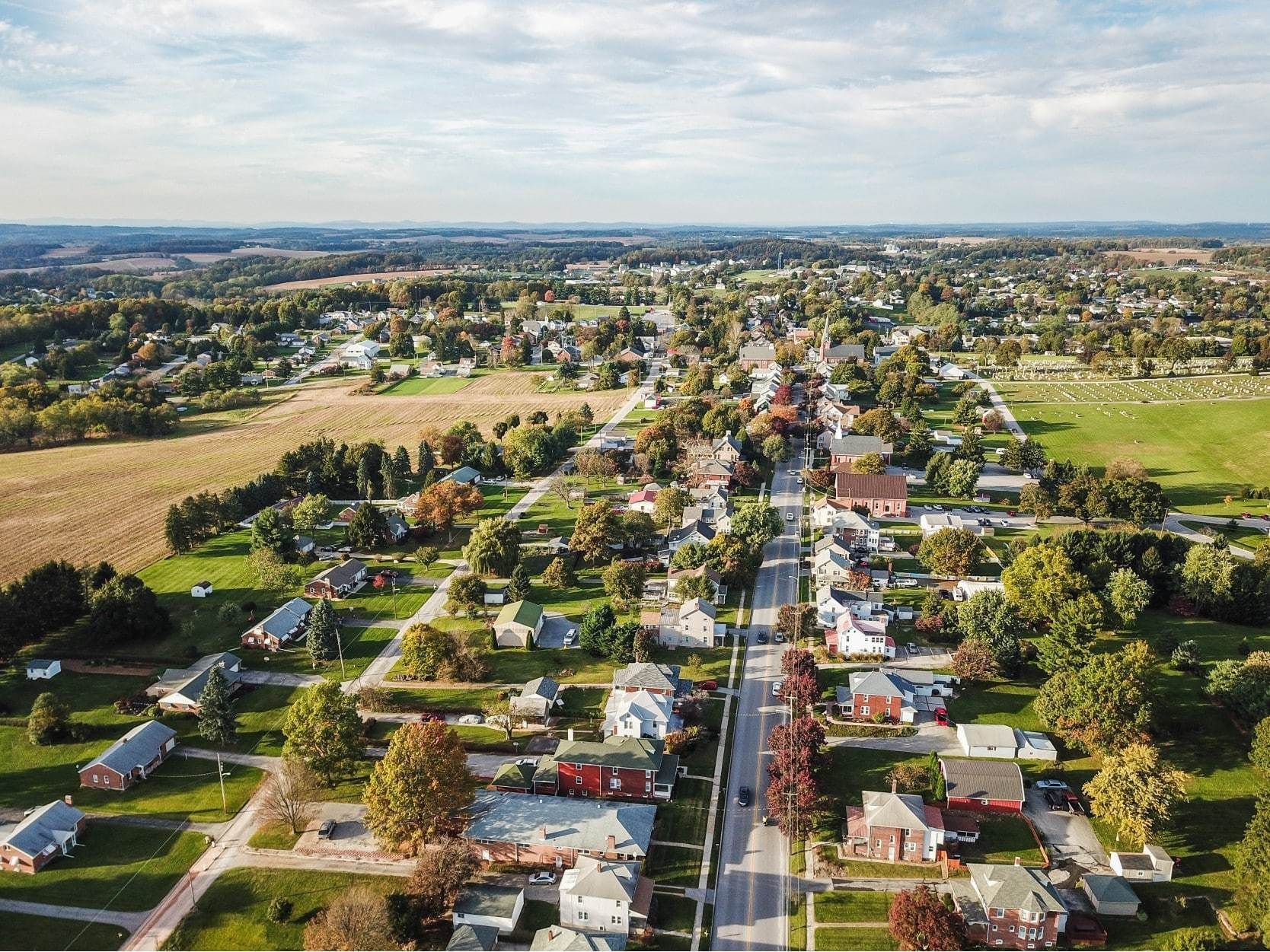 Aerial view of a small town with houses, trees, and a central road under a blue sky.