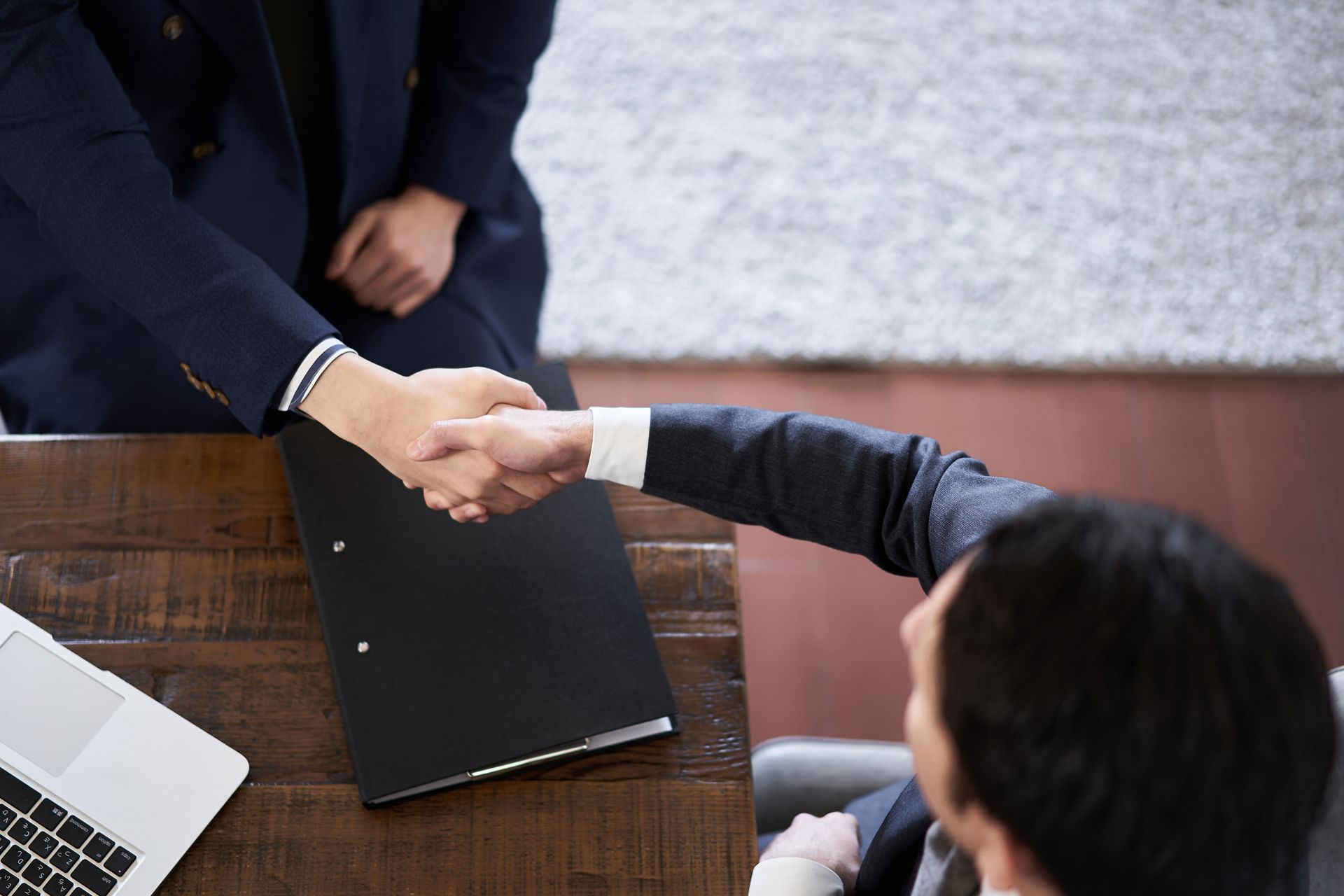 Two men in suits shaking hands over a wooden table with a laptop and a black folder.