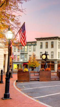 Streetscape with brick sidewalks, a clock tower, buildings, flags, and autumn foliage.