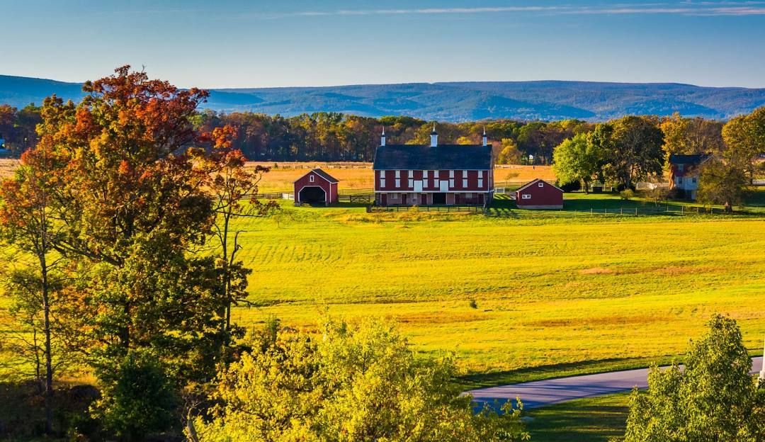 Scenic view: red-roofed house and outbuildings in a sunlit field, fall foliage, rolling hills in the background.