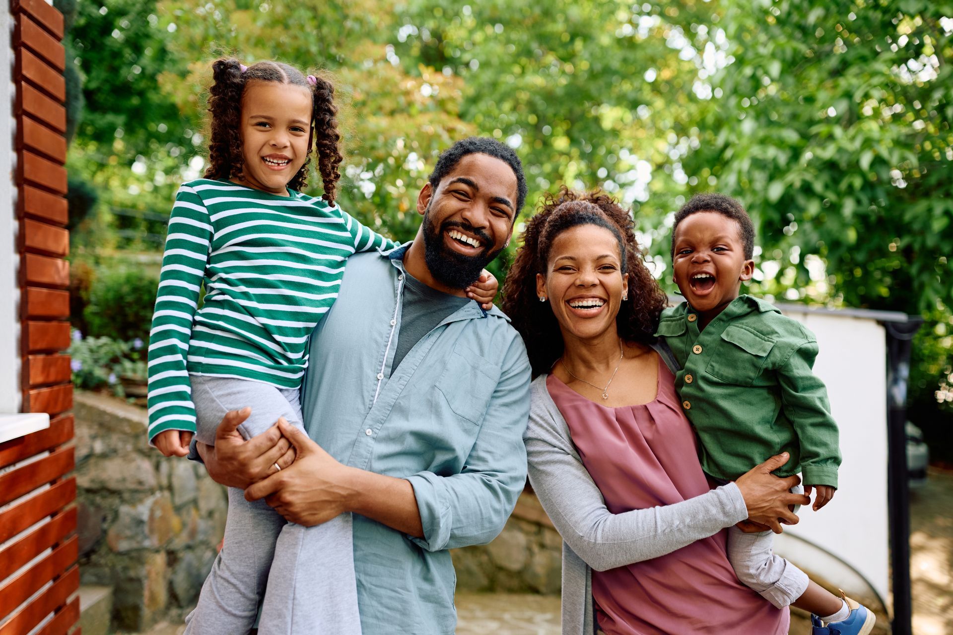 Family of four smiling outdoors, dad holding child, mom with another child, greenery in background.