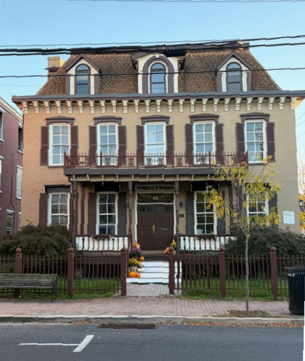 Brown and beige multi-story historic building with shutters, dormers, and a small front porch; iron fence.
