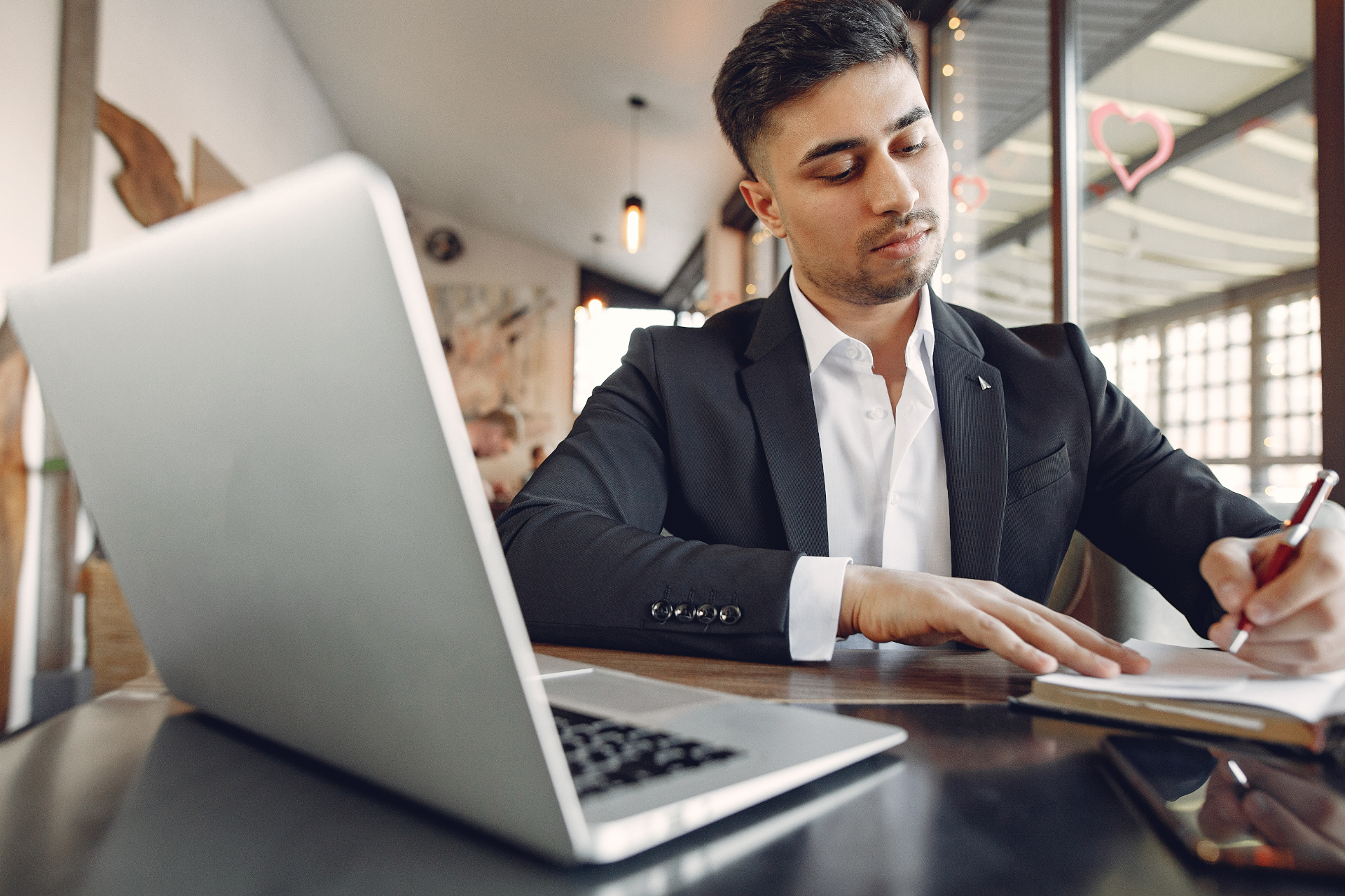 A person in a suit writing in a notebook at a table with a laptop in a modern, well-lit cafe setting.