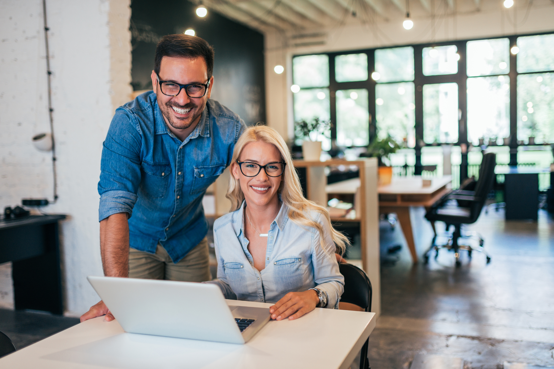 A man standing and a woman sitting at a desk with a laptop, both smiling, in a bright, modern office setting.