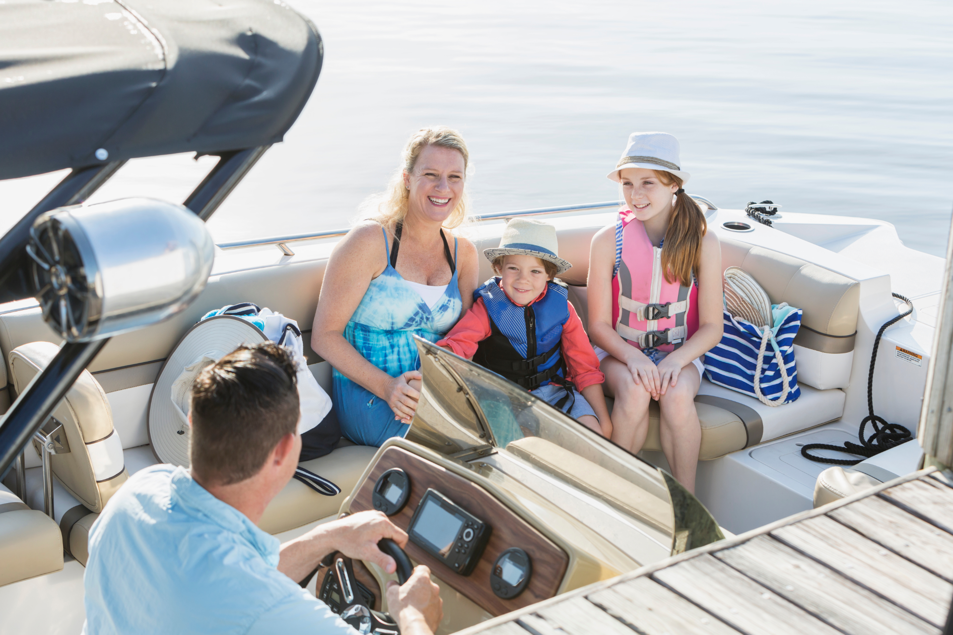 A family sits on a boat at a dock; a person steers while two others sit, and one stands near them, all smiling.