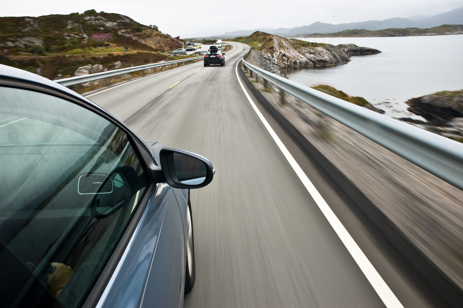 A car driving along a scenic coastal highway with rocky terrain and a body of water to the right.