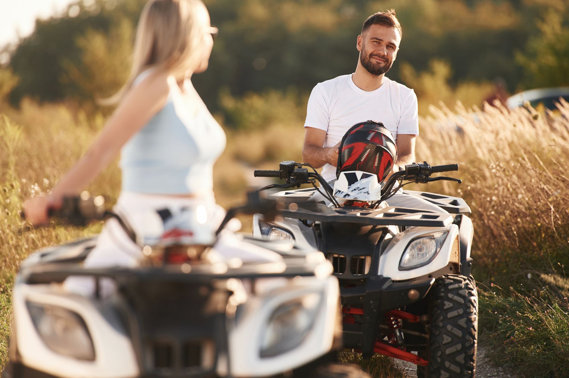 Two people riding white ATVs in a grassy field during a sunny day.
