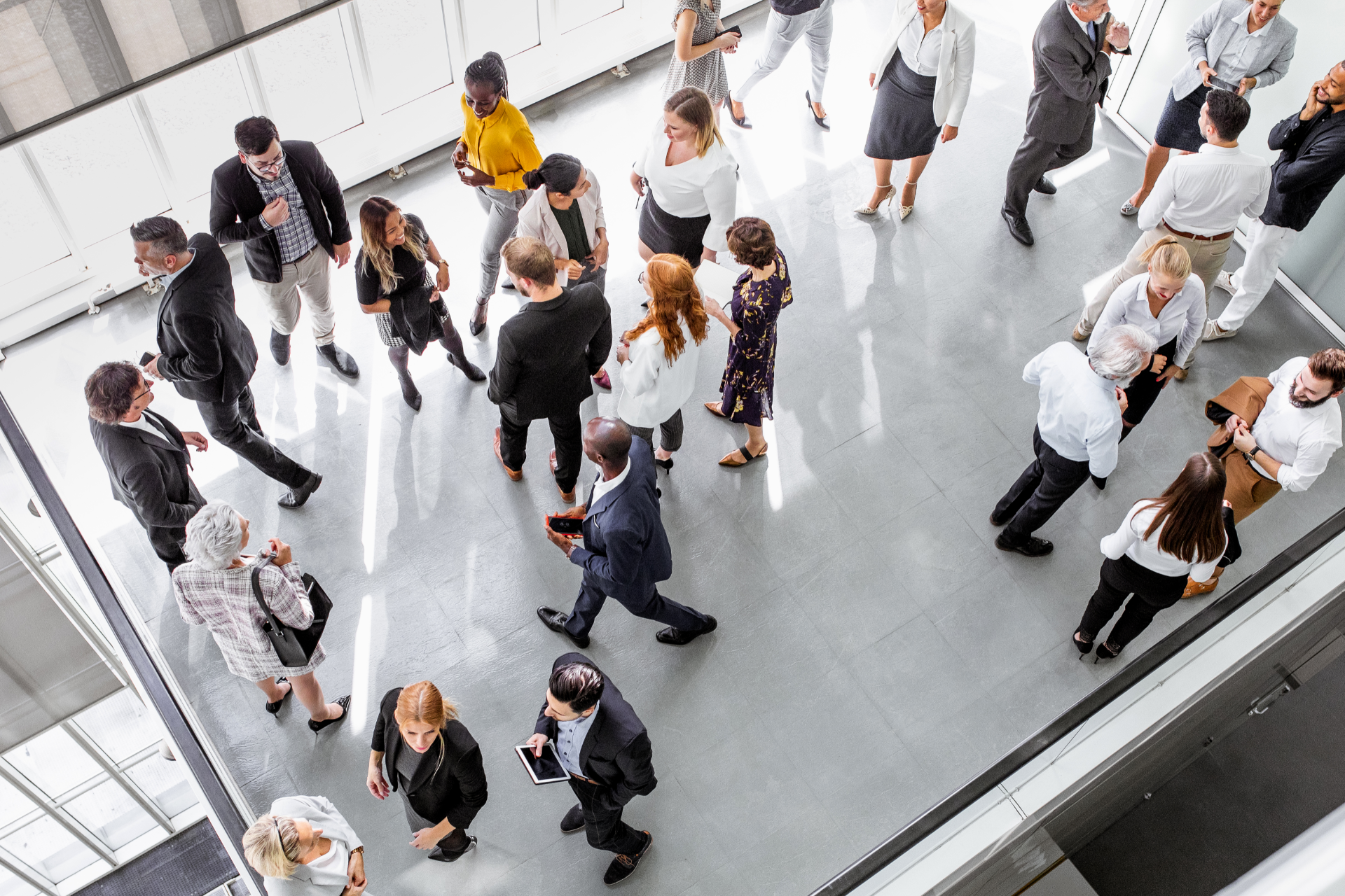 An elevated view of a diverse group of professionals in business attire mingling in a brightly lit, spacious modern lobby.