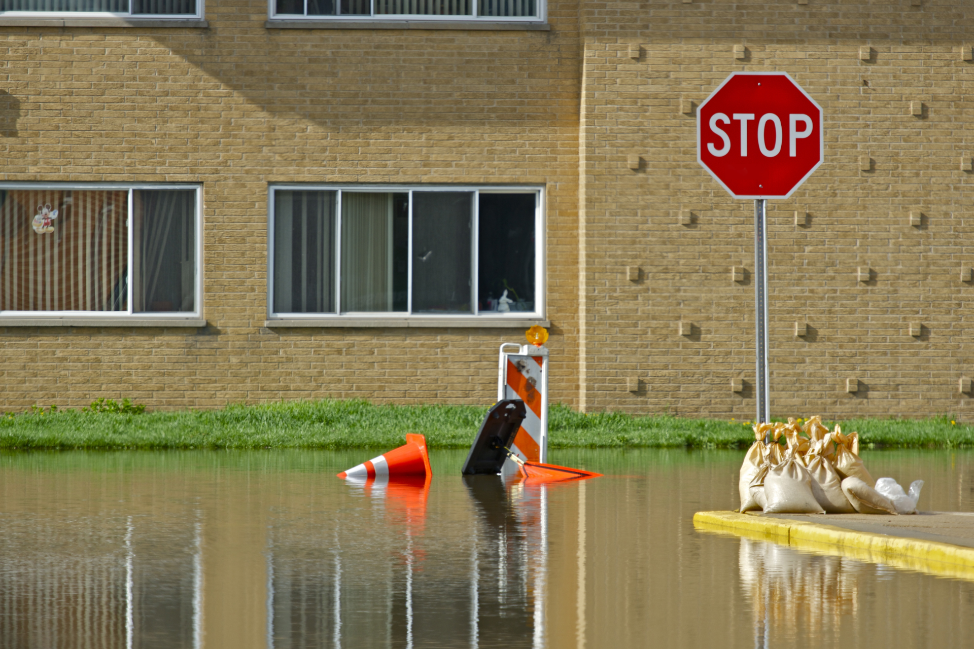 A flooded street in front of a brick building featuring a stop sign, traffic cones, and sandbags.